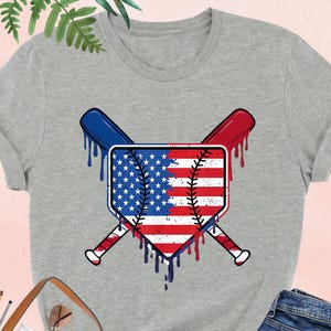 K&ouml;nnte beinhalten: Graues Athletic Heather T-Shirt mit Baseball-Motiv. Das Motiv zeigt ein Schild mit dem Design der amerikanischen Flagge, gekreuzte Baseballschl&auml;ger und tropfende Farbeffekte in Rot, Wei&szlig; und Blau. Das T-Shirt wird mit einer blauen Jeans gezeigt.