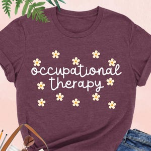 May include: A heather maroon t-shirt with the text "occupational therapy" in white script with small white flowers surrounding the text.