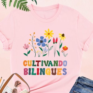 May include: Pink t-shirt with a floral design and the text "CULTIVANDO BILINGUES" in colorful letters. The shirt is paired with blue jeans, a brown tote bag, and black heels. The image also includes an umbrella, sunglasses, and a camera.