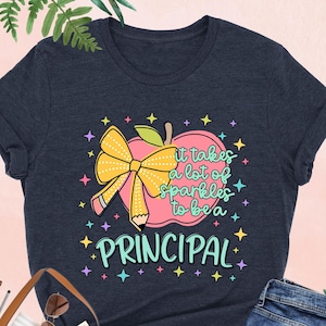 May include: A navy blue t-shirt with a colorful graphic that reads "It takes a lot of sparkles to be a Principal." The design includes an apple, pencil, and bow. The shirt is paired with blue jeans and a brown tote bag.