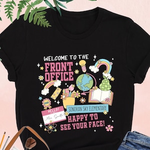 May include: Black t-shirt with a colorful graphic design featuring the text "Welcome to the Front Office", "Sonoran Sky Elementary", and "Happy to See Your Face!" The graphic also includes a globe, books, pencils, and flowers.