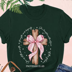 May include: A dark green t-shirt with a watercolor illustration of a pink ribbon tied around a wooden cross. The text "She laughs without fear" and "Proverbs 31:25" are printed on the shirt.