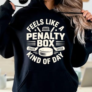 May include: Black hoodie with the text "FEELS LIKE A PENALTY BOX KIND OF DAY" in white. The design includes hockey sticks, a hockey puck, and a hockey net. The hoodie has a hood and a front pocket.