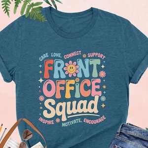 May include: A heather deep teal t-shirt with the words "FRONT OFFICE SQUAD" in colorful, retro-style lettering. The shirt includes the phrases "Care, Love, Connect, Support, Inspire, Motivate, Encourage". The shirt is paired with blue jeans and a brown tote bag.