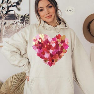 Pu&ograve; includere: Una felpa con cappuccio color sabbia con un grande disegno a cuore composto da pennellate rosa, rosse e dorate sovrapposte. La felpa con cappuccio ha un cappuccio con coulisse e una tasca frontale. La parola "SAND" &egrave; stampata in alto a destra.