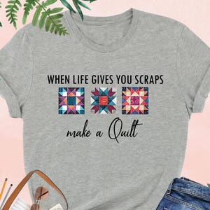 May include: Athletic heather gray t-shirt with the text "WHEN LIFE GIVES YOU SCRAPS make a Quilt." The shirt features three colorful quilt block designs. A brown leather tote bag, blue jeans, and black heels are also visible.