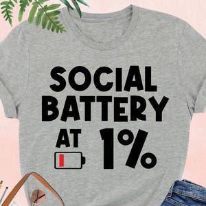 May include: Athletic heather gray t-shirt with the text "SOCIAL BATTERY AT 1%" in black. The design includes a low battery icon. The shirt is paired with blue jeans, a brown purse, and black heels.