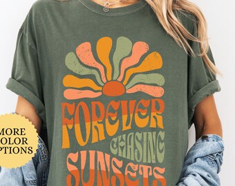 May include: A moss green t-shirt with a retro design. The graphic features a stylized sunset in orange, peach, and green, with the words "FOREVER CHASING SUNSETS" in orange and green. The shirt has rolled-up sleeves.
