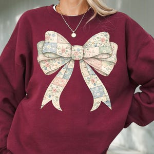 May include: Maroon sweatshirt with a large bow design. The bow features a patchwork pattern with floral and geometric elements in pastel colors. The sweatshirt has a crew neck and long sleeves.