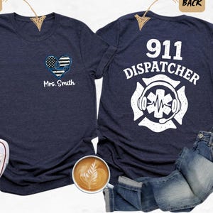 May include: A navy blue t-shirt with a white heart design featuring an American flag and the text "Mrs. Smith" on the front. The back of the shirt has a white design of a firefighter's helmet with a stethoscope and the text "911 Dispatcher".