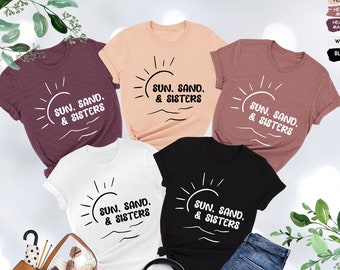 May include: Five t-shirts with a sun, sand, and sisters graphic design. The shirts are in various colors, including peach, mauve, maroon, white, and black.