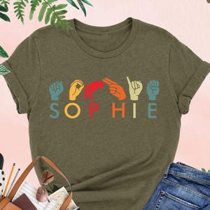 Puede incluir: Una camiseta verde con el nombre "SOPHIE" escrito en lenguaje de se&ntilde;as. Las letras est&aacute;n en un arco&iacute;ris de colores. La camiseta est&aacute; sobre una mesa con un bolso marr&oacute;n, unos vaqueros y un par de tacones negros.