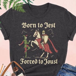Puede incluir: Camiseta de color gris oscuro con la inscripción "Born to Jest Forced to Joust" y un dibujo de un bufón, un caballero a caballo y un escudo. La camiseta se combina con unos vaqueros azules y un bolso tote marrón.