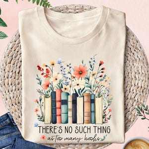 May include: Cream-colored t-shirt featuring a floral and book design. The design includes colorful flowers and a stack of books. The text "THERE'S NO SUCH THING as too many books" is printed below the books. The shirt is displayed on a woven mat.