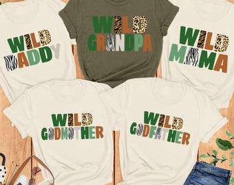 Könnte beinhalten: Fünf T-Shirts mit den Aufdrucken "Wild Daddy", "Wild Grandpa", "Wild Mama", "Wild Godmother" und "Wild Godfather". Der Text zeigt Tiermuster und grüne, braune und weiße Farben. Die T-Shirts liegen auf einer Holzoberfläche.