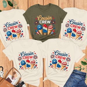 May include: Five t-shirts with the text "Cousin Crew" and illustrations of ice cream cones, popsicles, and fireworks. The shirts are in various colors, including white and olive green. A pair of blue jeans and a brown purse are also visible.