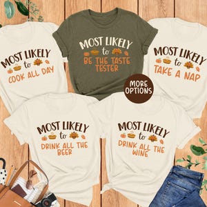 May include: Five t-shirts with fall-themed text. The shirts are cream and olive green, with phrases like "Most Likely to Cook All Day" and "Most Likely to Take a Nap" in brown. A pair of blue jeans and a brown purse are also in the image.