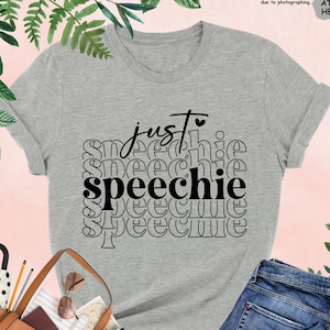 Puede incluir: Una camiseta gris jaspeada con el texto "just speechie" impreso en negro. El texto se repite varias veces en un patr&oacute;n apilado.