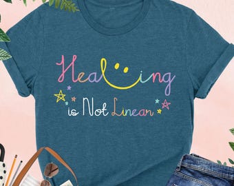 May include: A teal blue t-shirt with the text "Healing is Not Linear" in rainbow colors. The text includes a smiley face.