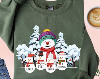 Op de afbeelding: Militairgroen sweatshirt met een sneeuwmanfamilie-afbeelding. De afbeelding bevat een sneeuwman met een paarse hoed met het woord "Oma" en vier kleinere sneeuwmannen met de namen: Emily, Mike, Sarah en Thomas. De sweatshirt heeft lange mouwen.