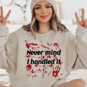 Puede incluir: Sudadera de color arena con el texto "Never mind I handled it" en negro, con salpicaduras rojas y gráficos de huellas de manos. La modelo lleva un gorro a juego. La sudadera es de estilo casual y cómodo.