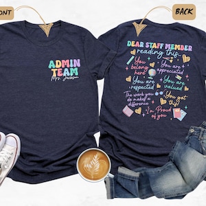 May include: Heather navy t-shirts with text. The front shirt says "ADMIN TEAM Mrs. Jackson" in colorful letters. The back shirt says "DEAR STAFF MEMBER reading this..." with additional text and colorful graphics. A pair of white sneakers, jeans, and a coffee cup are also in the image.