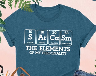 May include: A teal blue t-shirt with a white graphic of the periodic table elements, with the text "The Elements of My Personality" underneath. The elements shown are Sulfur (S), Argon (Ar), Calcium (Ca), and Samarium (Sm).