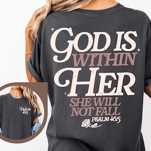 May include: Dark gray t-shirt with the text "God Is Within Her" in white and mauve lettering. The back of the shirt also features the text "She Will Not Fall" and "Psalm 46:5". A small graphic of a rose is below the text.