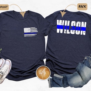 May include: A navy blue t-shirt with a thin blue line American flag on the front pocket and the text "WILSON" with a thin blue line above it on the back.