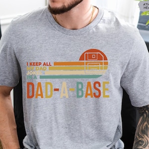 May include: A gray t-shirt with a retro-style graphic that says "I keep all my dad jokes in a dad-a-base". The graphic features a floppy disk icon and colorful stripes.