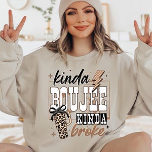 Puede incluir: Sudadera color arena con el texto "kinda BOUJEE kinda broke" en una mezcla de fuentes y un gráfico de vaso con estampado de leopardo. La sudadera tiene mangas largas y cuello redondo. La persona lleva un gorro.