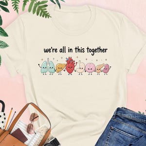 May include: A natural-colored t-shirt featuring a graphic of cartoon human organs holding hands with the text "we're all in this together" printed above them. The organs include lungs, liver, heart, stomach, brain, pancreas, and kidneys.