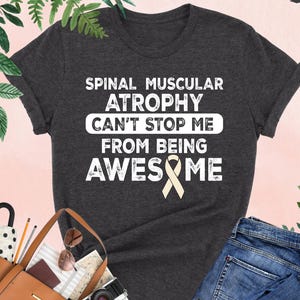 May include: A dark grey heather t-shirt featuring the white text SPINAL MUSCULAR ATROPHY CAN'T STOP ME FROM BEING AWESOME with a light beige awareness ribbon integrated into the word AWESOME. The shirt has short sleeves and a crew neckline.