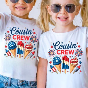 May include: Two children wearing white t-shirts with the text "Cousin Crew" in blue script. The shirts feature patriotic ice cream cone and firework graphics in red, white, and blue. The children are wearing sunglasses.