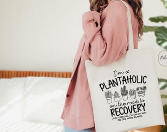 May include: White tote bag with black text that reads "I'm a PlantahOLIC on the road to RECOVERY. Just kidding, I'm on my way to get more plants."  The bag features a graphic of potted plants.