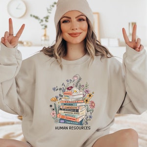 May include: A light beige sweatshirt with a graphic design of a stack of books with flowers. The books have text on the spines that reads "Inclusion", "Compassion", "Ethical", "Professional Growth", "Advocacy", "Motivational", and "Team Building". The text "Human Resources" is printed below the graphic.