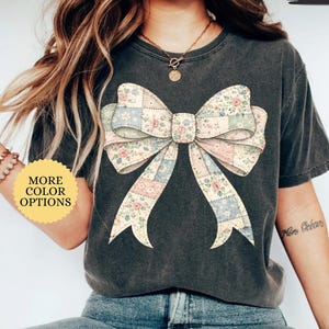 May include: Dark gray t-shirt featuring a large bow design with a patchwork pattern of floral and pastel squares. The shirt has the word "PEPPER" printed on the top right. A button on the left says "MORE COLOR OPTIONS".