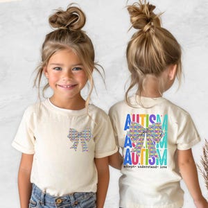 May include: Two children wearing cream-colored t-shirts with a colorful bow graphic. The front of the shirt has a bow graphic, and the back has the word "AUTISM" in blue, green, and yellow, with the words "accept- understand- love".