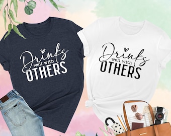May include: Two t-shirts with the text "Drinks well with OTHERS" in black lettering on a white background and a heather navy background.