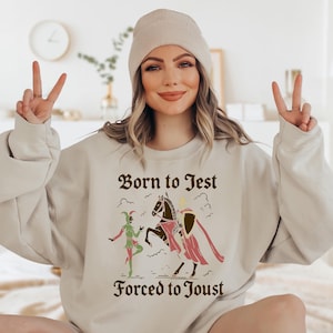 May include: Sand-colored sweatshirt with the text "Born to Jest, Forced to Joust" in a medieval font. The graphic features a jester, a knight on horseback, and decorative elements. The model is wearing a beige beanie.