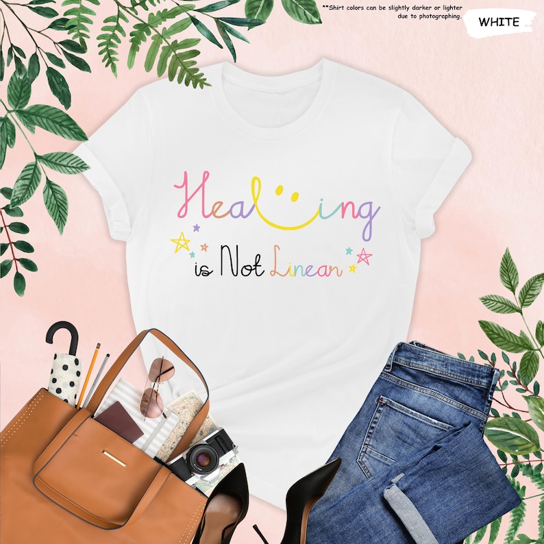 K&ouml;nnte beinhalten: Wei&szlig;es T-Shirt mit dem Text "Healing is Not Linear" in Regenbogenfarben mit einem Smiley und Sternen.