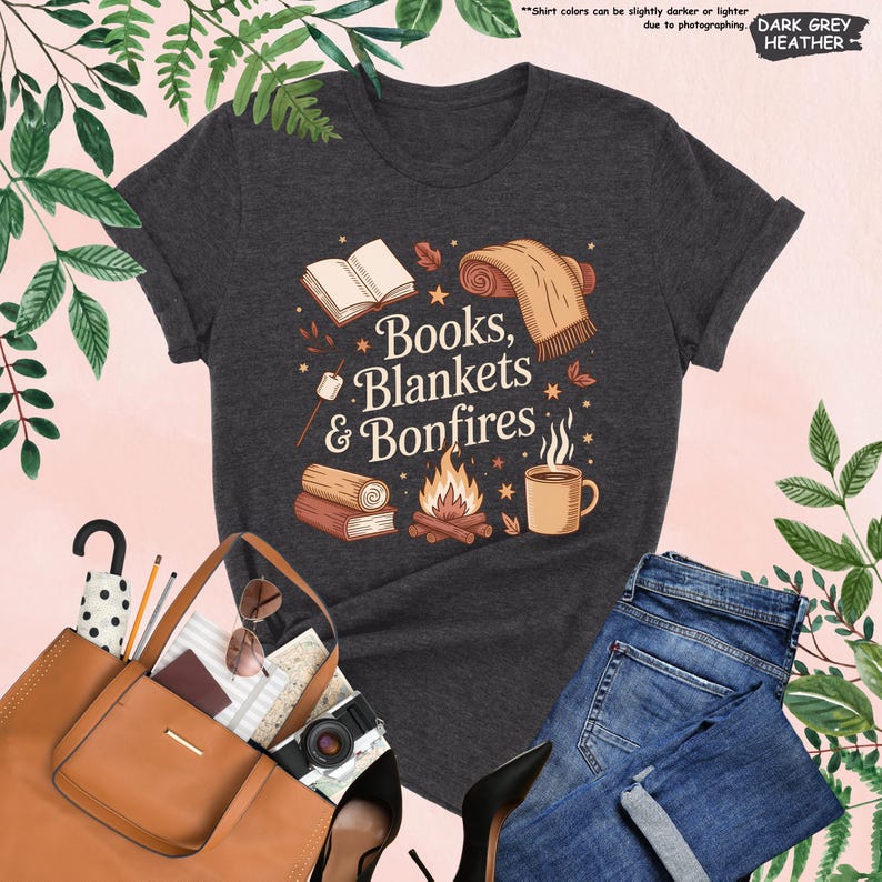May include: Dark grey heather t-shirt with the text "Books, Blankets & Bonfires" and illustrations of a book, blanket, campfire, and mug. The shirt is paired with blue jeans, a brown tote bag, and black heels.