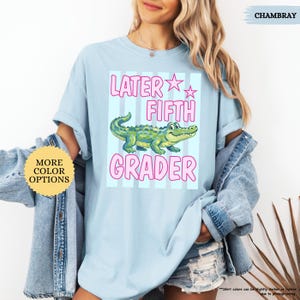 May include: Light blue t-shirt with a graphic of a green alligator and the text "LATER FIFTH GRADER" in pink. The shirt has short sleeves and a crew neck. The words "MORE COLOR OPTIONS" are on a yellow circle.