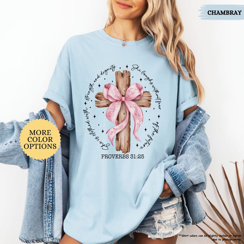 Op de afbeelding: Lichtblauw T-shirt met een houten kruis en een roze strik. Het kruis heeft de tekst "Zij is bekleed met kracht en waardigheid. Zij lacht zonder angst voor de toekomst." en "Spreuken 31:25" erop gedrukt.