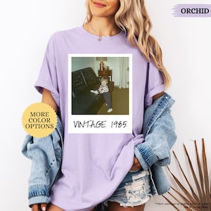 Op de afbeelding: Een paars T-shirt met een vintage Polaroid-foto van een klein kind dat op een bank zit. De foto is gelabeld met "Vintage 1985".