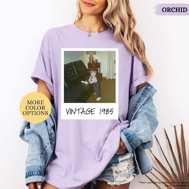 Op de afbeelding: Een paars T-shirt met een vintage Polaroid-foto van een klein kind dat op een bank zit. De foto is gelabeld met "Vintage 1985".