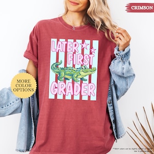 May include: Crimson-colored t-shirt with the text "LATER FIRST GRADER" in pink and a cartoon alligator. The design is set against a light blue striped background. The shirt has rolled-up sleeves and a crew neck.