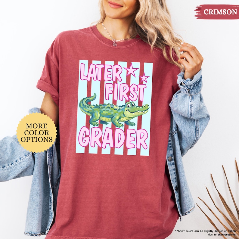 May include: Crimson-colored t-shirt with the text "LATER FIRST GRADER" in pink and a cartoon alligator. The design is set against a light blue striped background. The shirt has rolled-up sleeves and a crew neck.