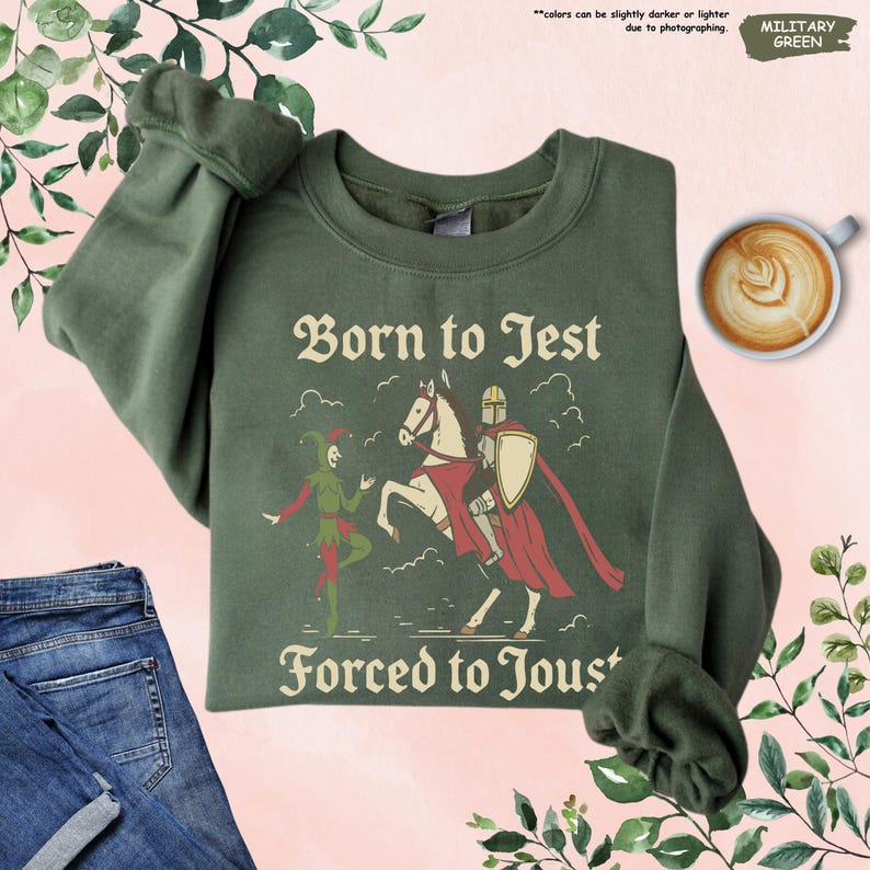 K&ouml;nnte beinhalten: Milit&auml;rgr&uuml;nes Sweatshirt mit dem Text "Born to Jest, Forced to Joust" und einer Grafik eines Narren, Ritters und Pferdes. Das Sweatshirt hat einen Rundhalsausschnitt und lange &Auml;rmel. Eine Tasse Kaffee und eine Jeans sind im Hintergrund.