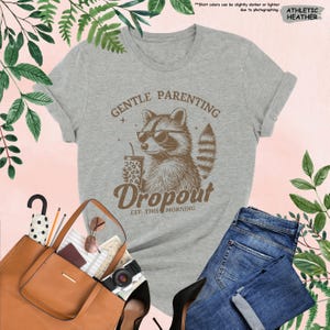 May include: Athletic heather gray t-shirt with a raccoon wearing sunglasses and holding a drink. The shirt has the words "GENTLE PARENTING Dropout EST. THIS MORNING" printed on it. The shirt is paired with blue jeans, a brown tote bag, and black heels.